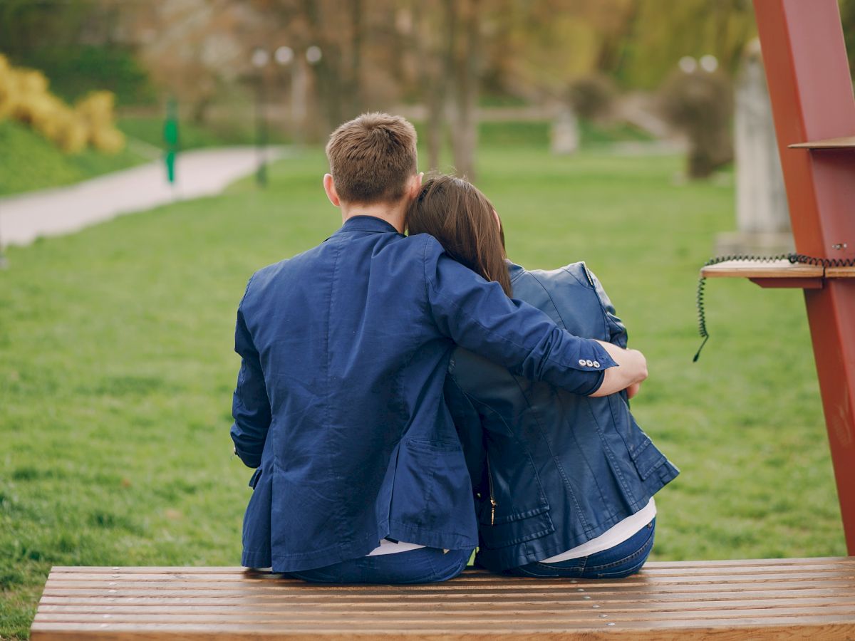 Two people are sitting on a bench outside, with one's arm around the other. They are wearing jackets and appear to be embraced.