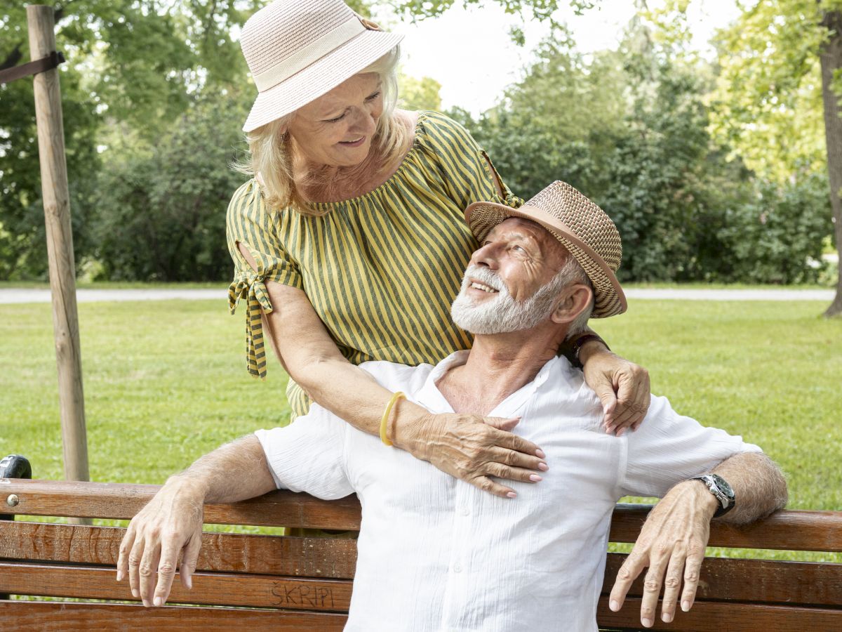 An elderly couple, wearing hats, shares a moment on a park bench, the woman standing behind the seated man, smiling affectionately.