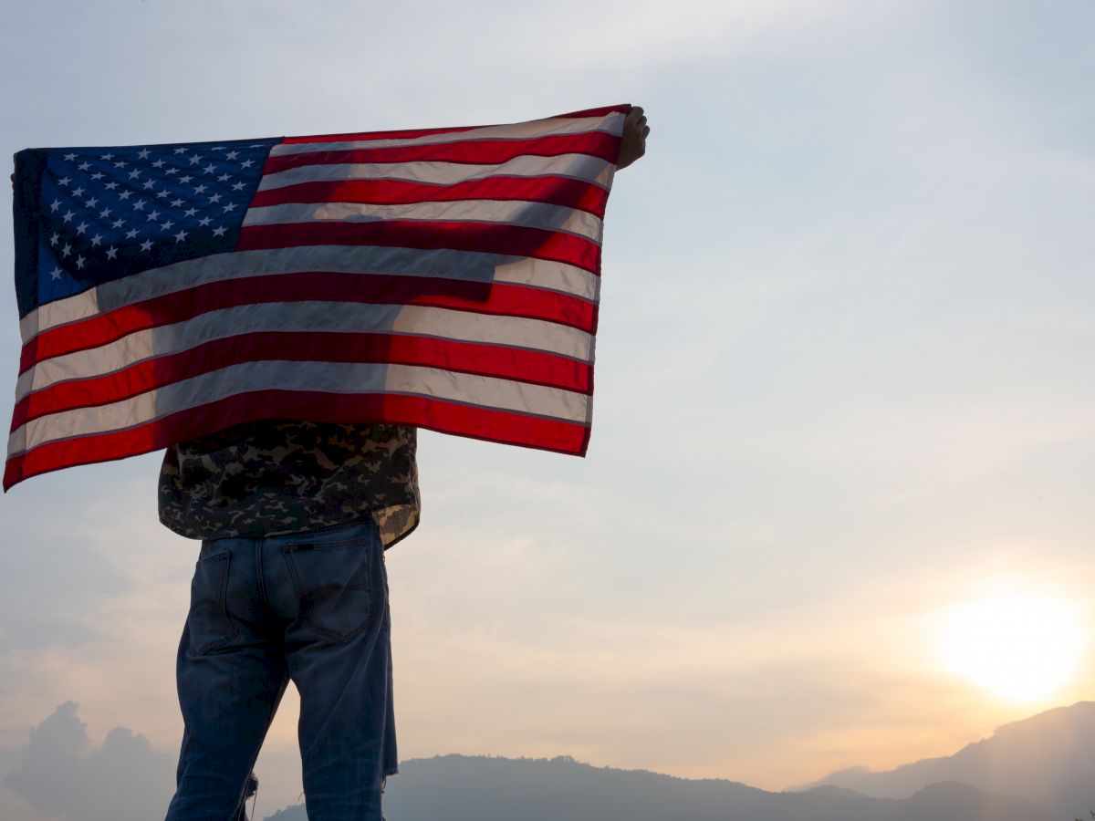 Person holding up an American flag against the backdrop of mountains and a setting sun, creating a silhouette.