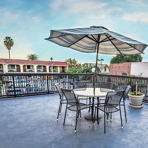 A patio with a table, four chairs, and an umbrella overlooks a view of a building and palm trees under a clear sky, ending the sentence.