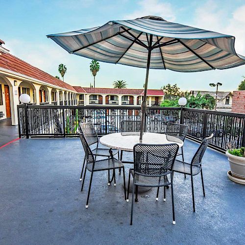 An outdoor patio area with a table, four chairs, and a large umbrella, overlooking a courtyard with palm trees and buildings in the background.