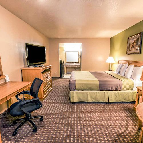 A clean, well-lit hotel room with a king-sized bed, TV, desk, and seating area. There are two lamps and a framed picture on the wall.