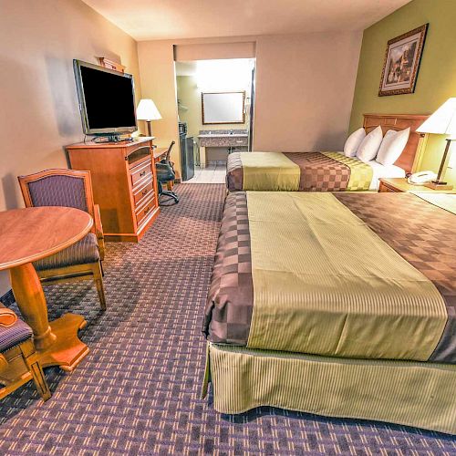 This image shows a hotel room with two double beds, a TV, a table with chairs, and a view into the bathroom area in the background.