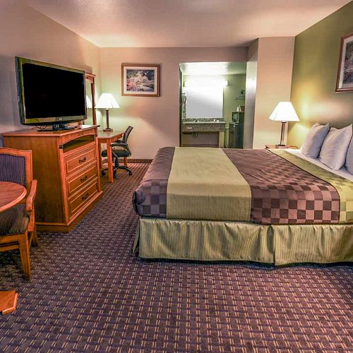 The image shows a hotel room with a king-sized bed, a TV, a round table with chairs, a dresser, two lamps, and decorated walls with framed pictures.