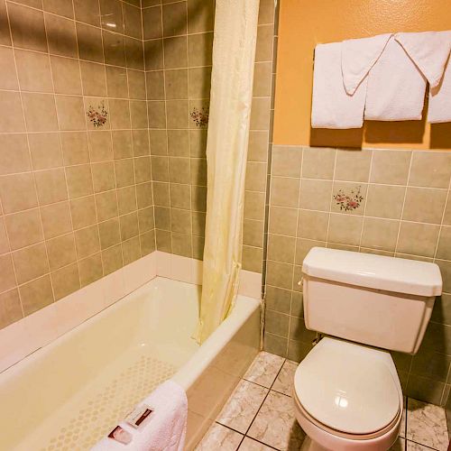 A bathroom with a green-tiled bathtub, beige shower curtain, white toilet, and folded towels hanging on the wall.