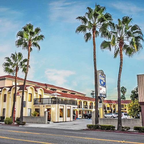 The image shows a two-story motel with an exterior corridor, palm trees, and a sign. The sky is partly cloudy and the street is visible in the foreground.