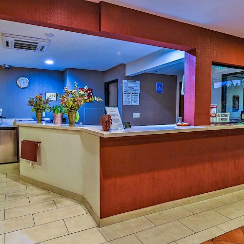 The image shows a hotel reception area with a counter, flower arrangements, a clock, and some informational signs on the walls.