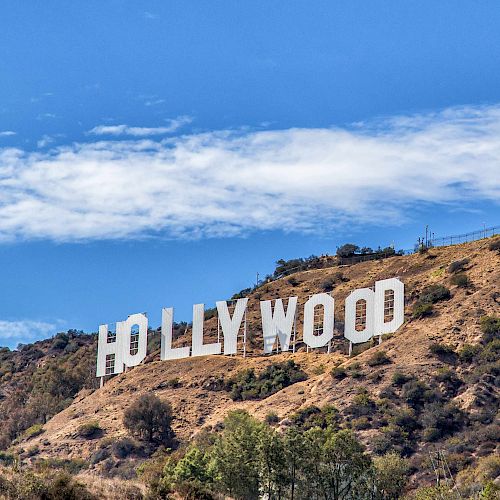 The image shows the iconic Hollywood sign located in the hills of Los Angeles, California, under a bright blue sky with some clouds.