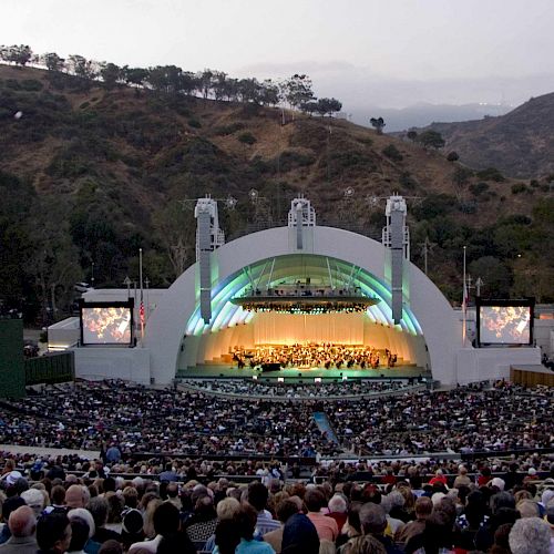 A large crowd is gathered at an outdoor amphitheater surrounded by hills, watching a performance on a brightly lit stage.