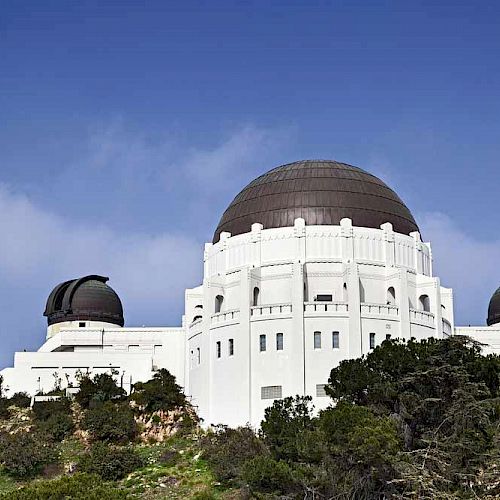 The image shows a large white observatory with multiple domes situated atop a hill, surrounded by greenery and under a clear blue sky.