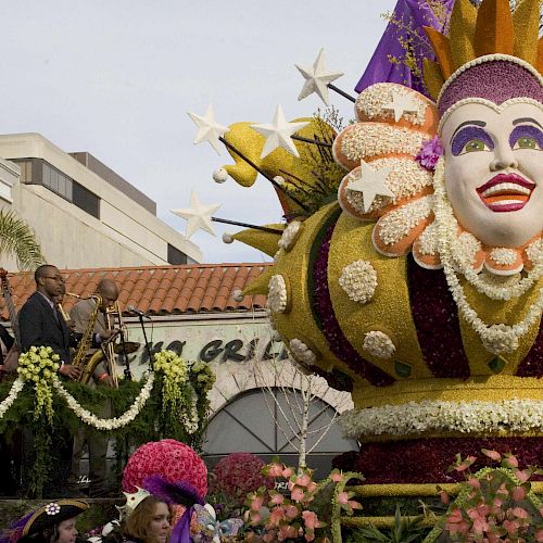 The image shows a festive parade float with a large colorful mask surrounded by flowers, and people observing it from a decorated platform.