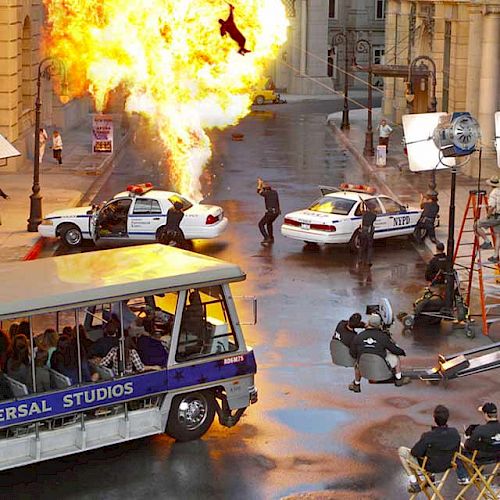 A film set at Universal Studios with an explosion scene, police cars, crew, equipment, and a tour tram with onlookers in the foreground.