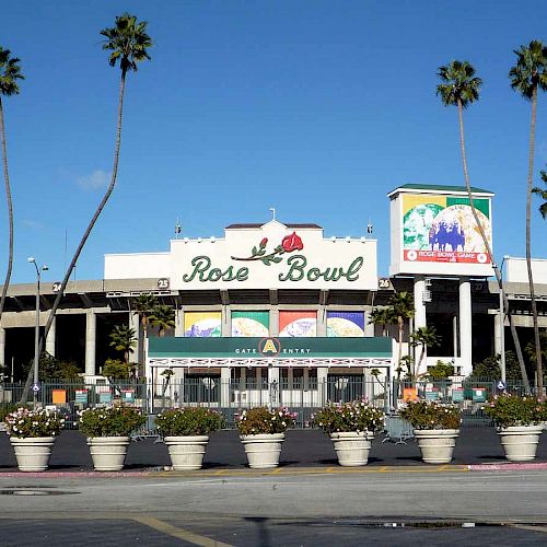 An exterior view of the Rose Bowl stadium surrounded by palm trees and flower pots on a sunny day, with colorful banners displayed on the building.