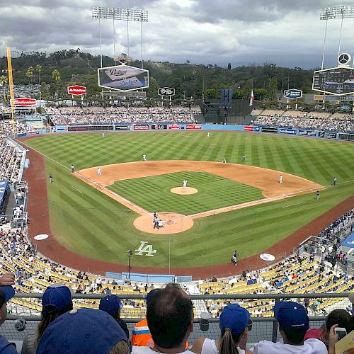 A baseball stadium filled with fans watching a game from the stands, with players positioned on the field and vibrant green grass under a cloudy sky.