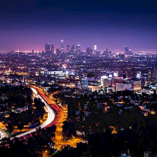 A cityscape at night shows a sprawling urban area with a brightly lit skyline and a curving road with streaks of car lights.