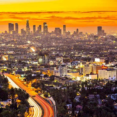 A vibrant cityscape with tall buildings under an orange sunset sky, showcasing a busy highway with light trails of moving vehicles.