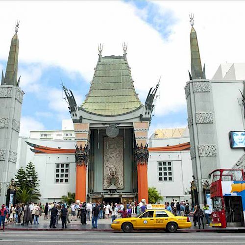 A busy street scene featuring a tall, elaborate building entrance with decorative towers, a yellow taxi, and a double-decker bus with 