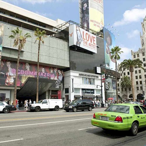 A busy street scene with various cars, including a green taxi, billboards advertising shows, and buildings with palm trees lining the street.