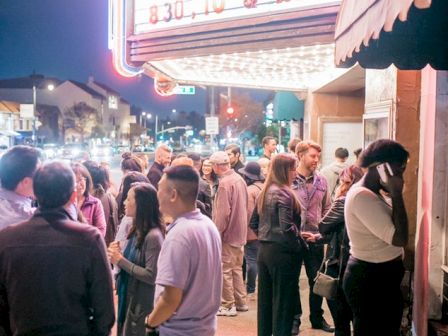 A crowd of people stands outside a theater with a marquee displaying showtimes at night, illuminated by bright neon lights.