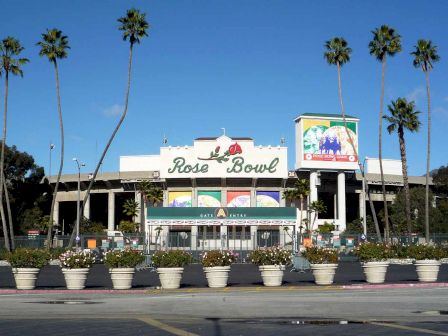 This image shows the exterior facade of the Rose Bowl stadium, with palm trees and flower pots in the foreground against a clear blue sky.