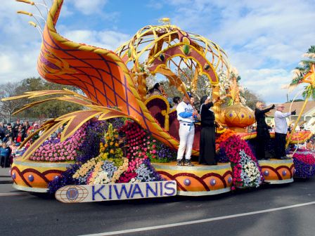 A colorful parade float adorned with flowers and 'KIWANIS' signs, featuring performers in costumes, and a crowd watching in the background.