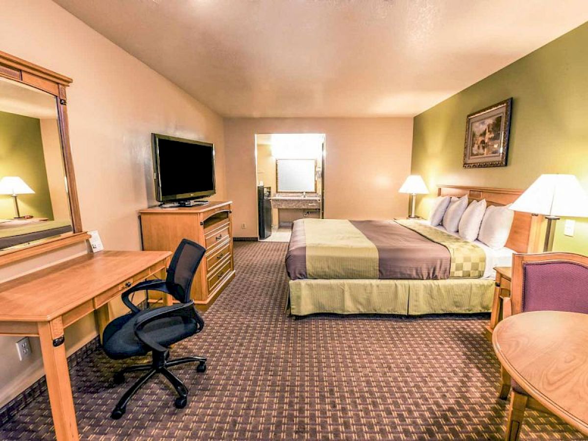 The image is of a hotel room with a king-sized bed, desk, chair, TV, lamps, a mirror, and a small table with chairs. The room is warmly lit.