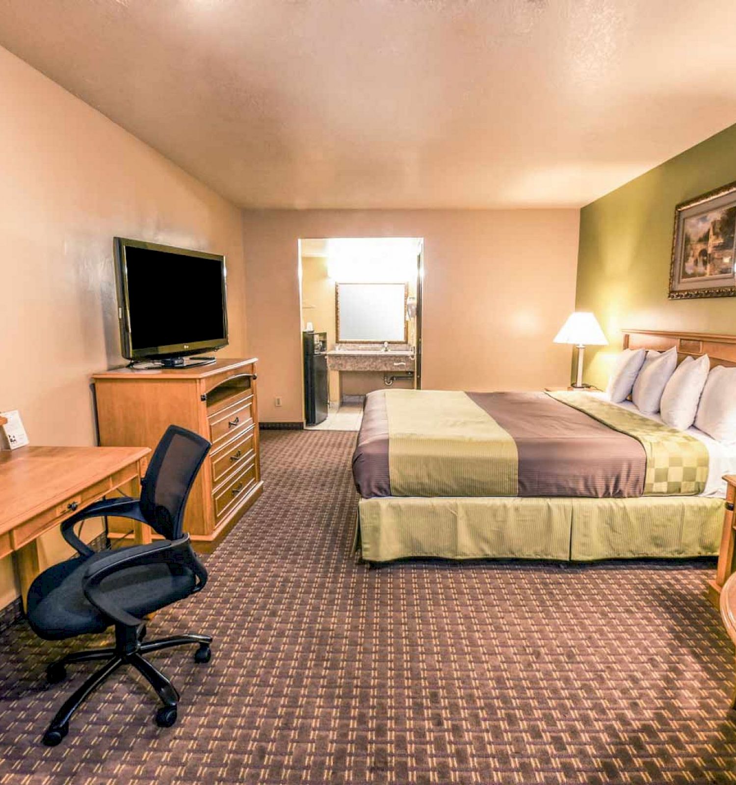 A hotel room with a large bed, TV, desk with chair, two lamps, and a small table with chairs. The walls are beige and green.