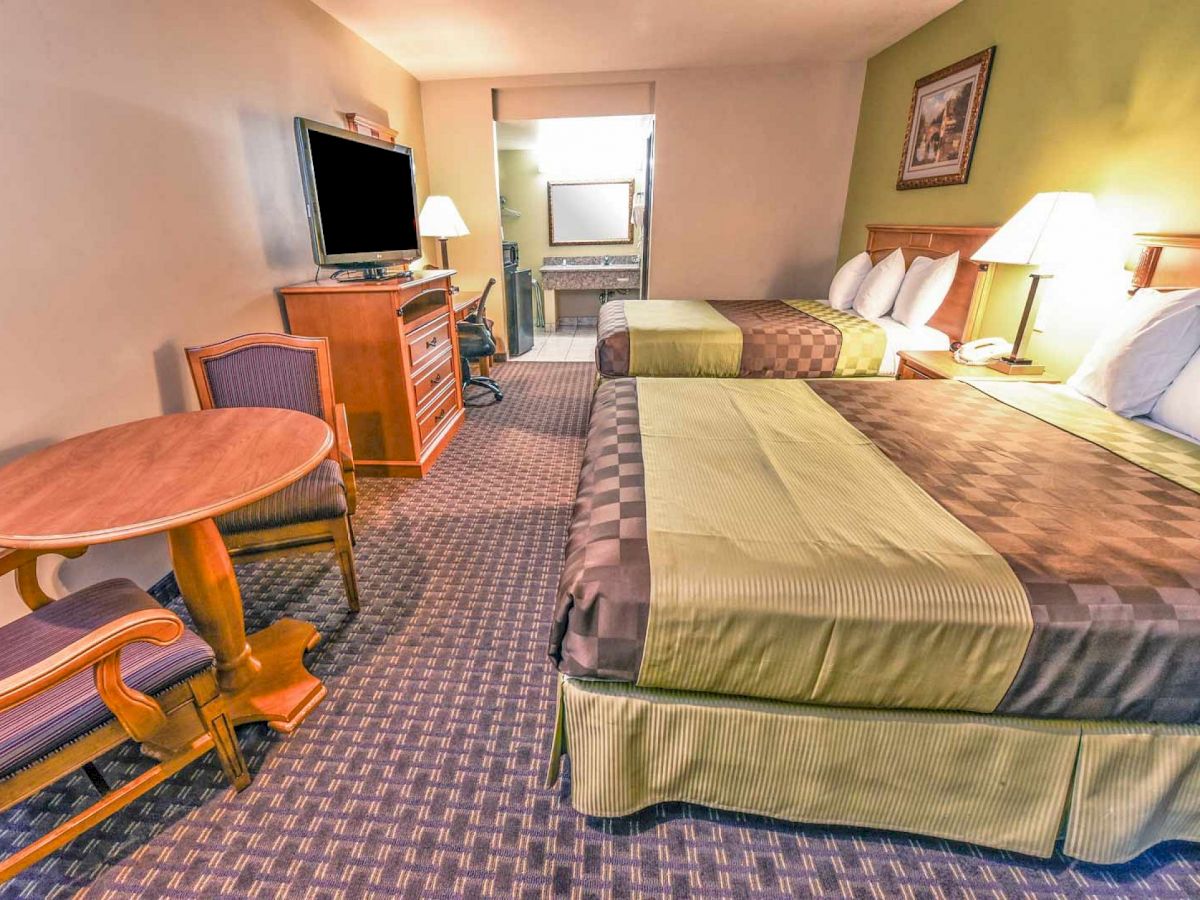 The image shows a hotel room with two double beds, a flat-screen TV, a dresser, a small round table with two chairs, and a view into a bathroom.