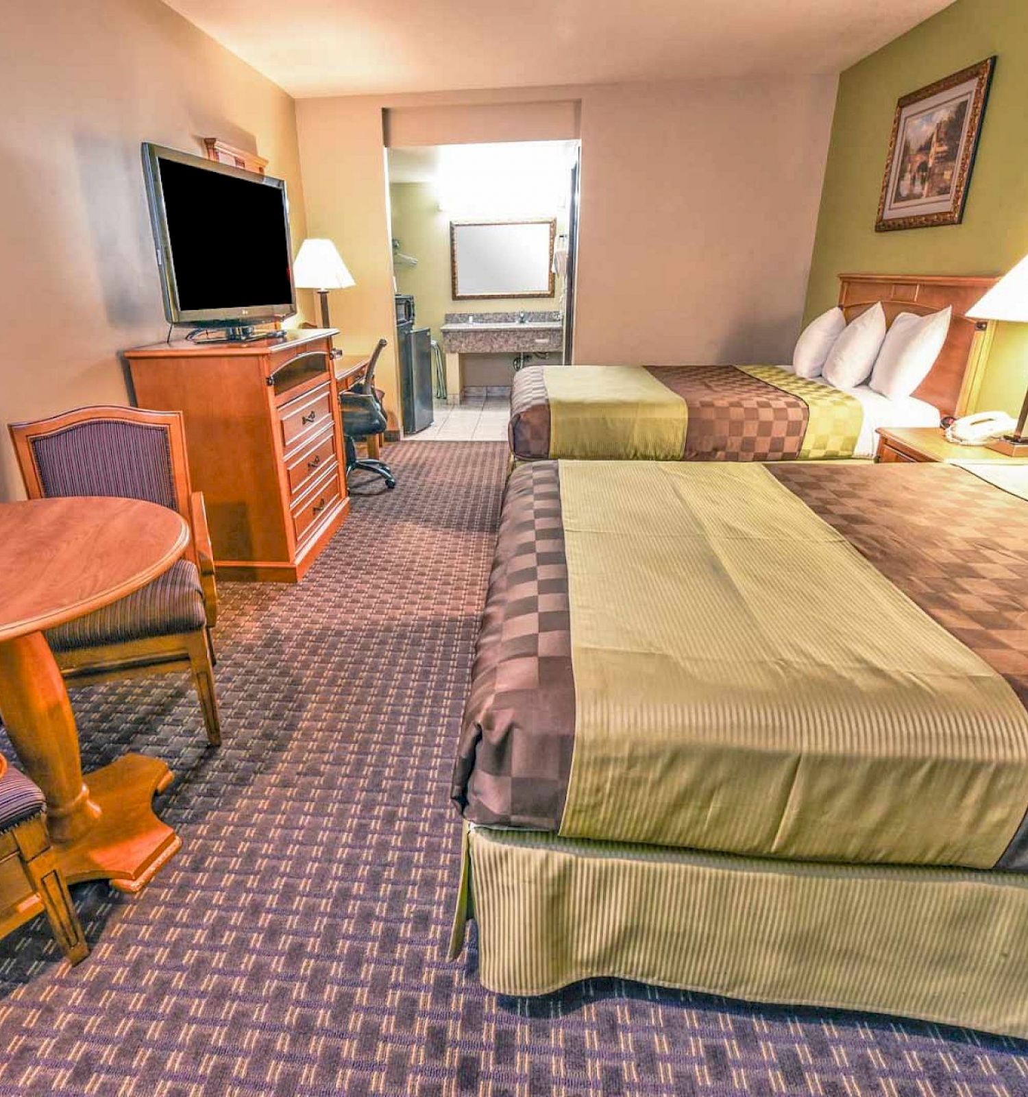 The image shows a hotel room with two double beds, a flat-screen TV, a dresser, a small round table with two chairs, and a view into a bathroom.