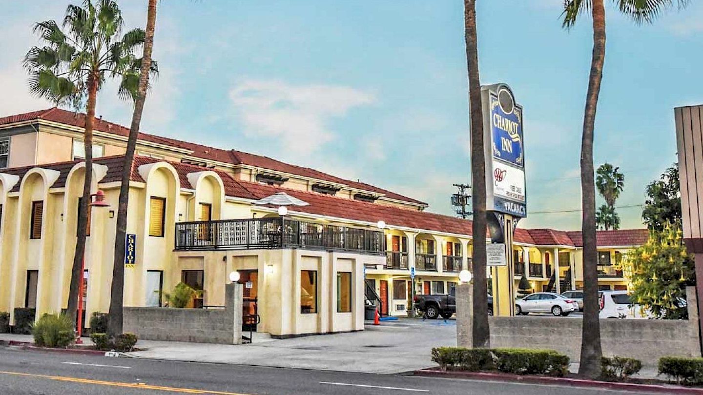 The image shows a motel with a "Quality Inn" sign, palm trees, and some vehicles parked in the lot. Text in a foreign language is visible.