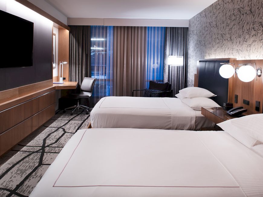 The image shows a modern hotel room with two beds, a large TV, a desk and chair by the window, and contemporary decor.