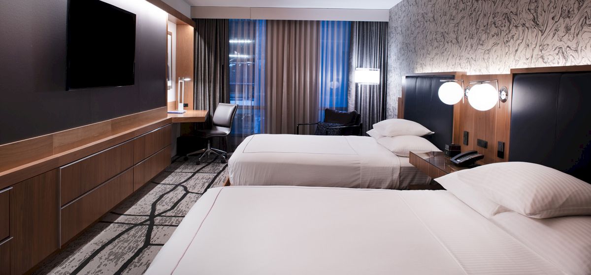 The image shows a modern hotel room with two beds, a large TV, a desk and chair by the window, and contemporary decor.
