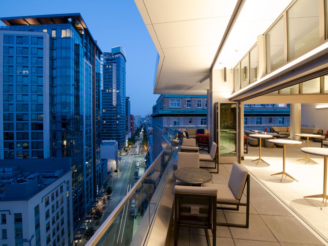 The image shows a modern cityscape from a high balcony during the evening, with buildings, a street view, outdoor seating, and indoor spaces.