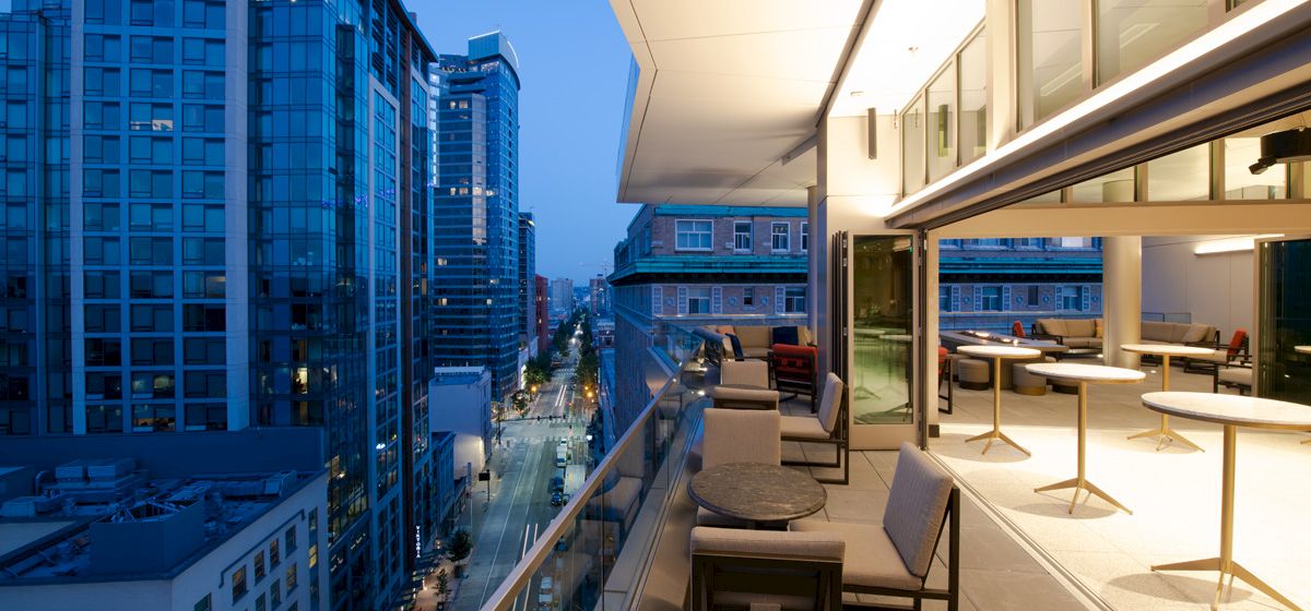 The image shows a modern cityscape from a high balcony during the evening, with buildings, a street view, outdoor seating, and indoor spaces.