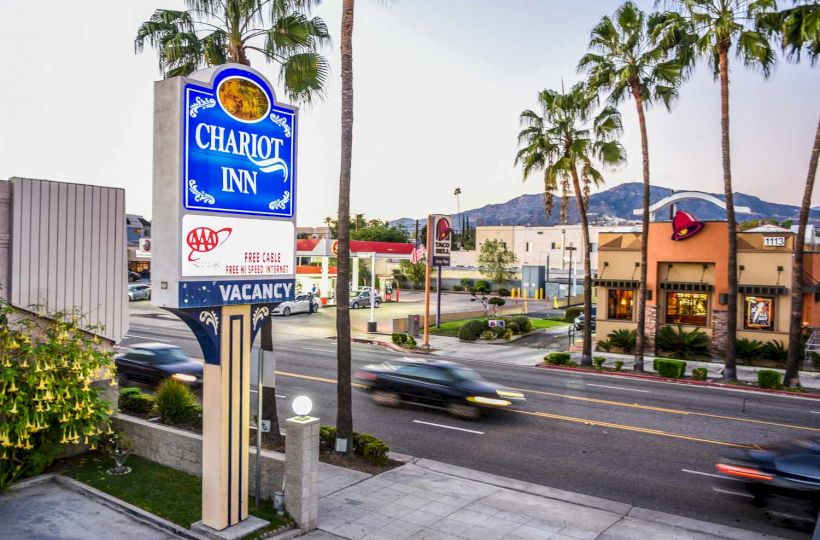 A street view showing the Chariot Inn sign, palm trees, nearby businesses, and a road with moving cars under a clear sky.