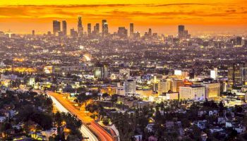 A vibrant cityscape at sunset with a glowing orange sky, featuring skyscrapers and a bustling highway with light trails from moving vehicles.