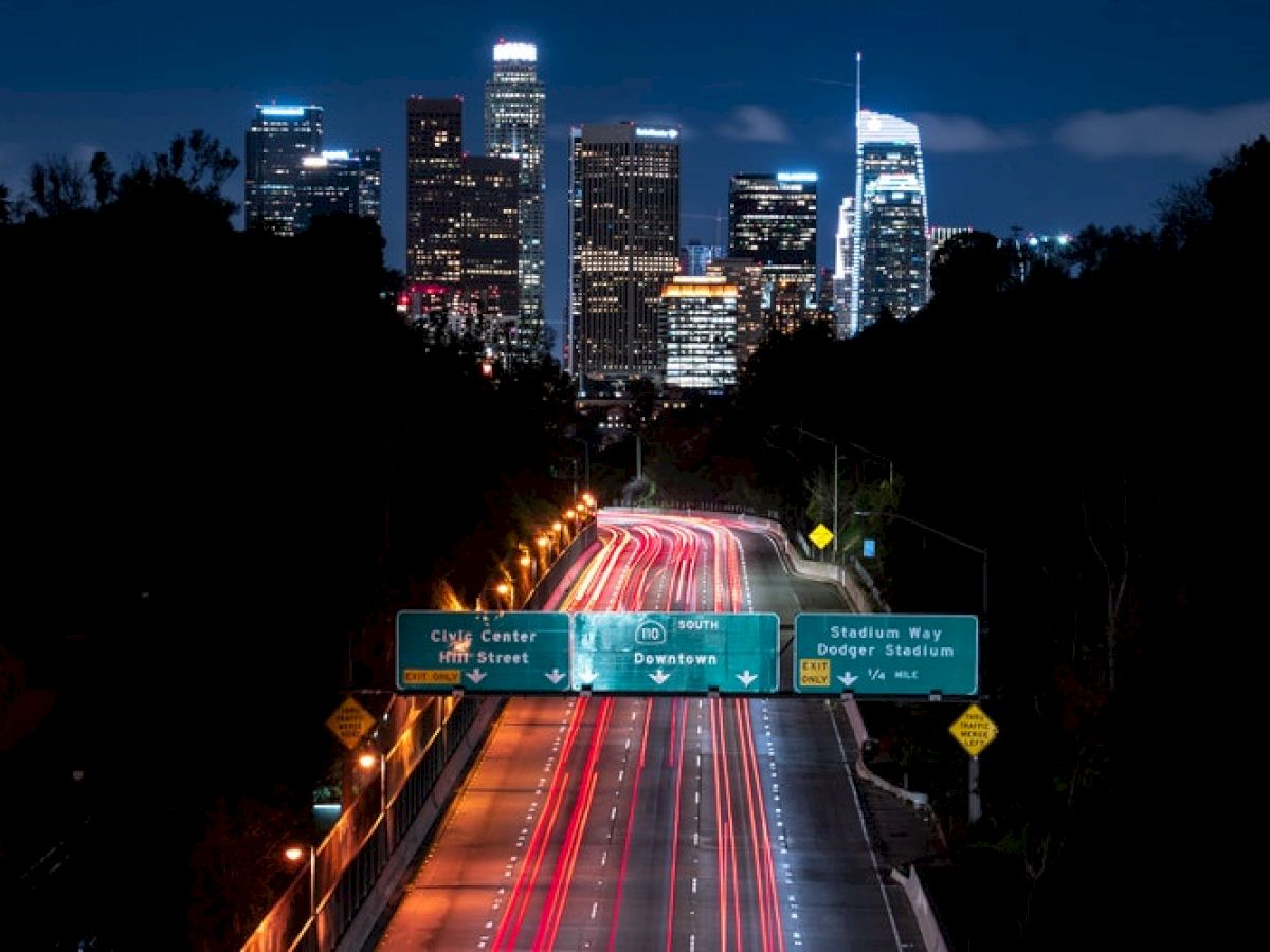 The image shows a nighttime cityscape with a freeway leading towards a skyline of illuminated buildings, featuring road signs for City Center and Stadium Way.