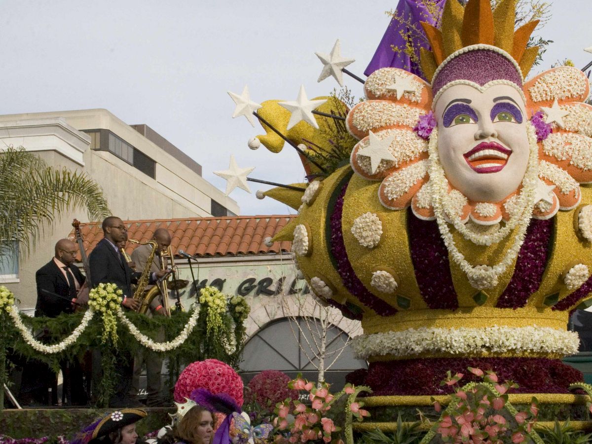 The image shows a colorful parade float featuring a large, ornate mask. People are on the stage decorated with flowers near the float.