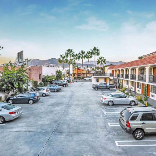 A parking lot with several cars is adjacent to a two-story motel surrounded by palm trees and mountain views in the background.