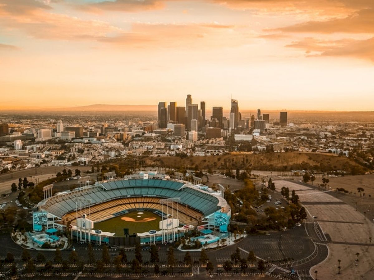 An aerial view shows a stadium with a baseball field in the foreground and a city skyline in the background during sunset.