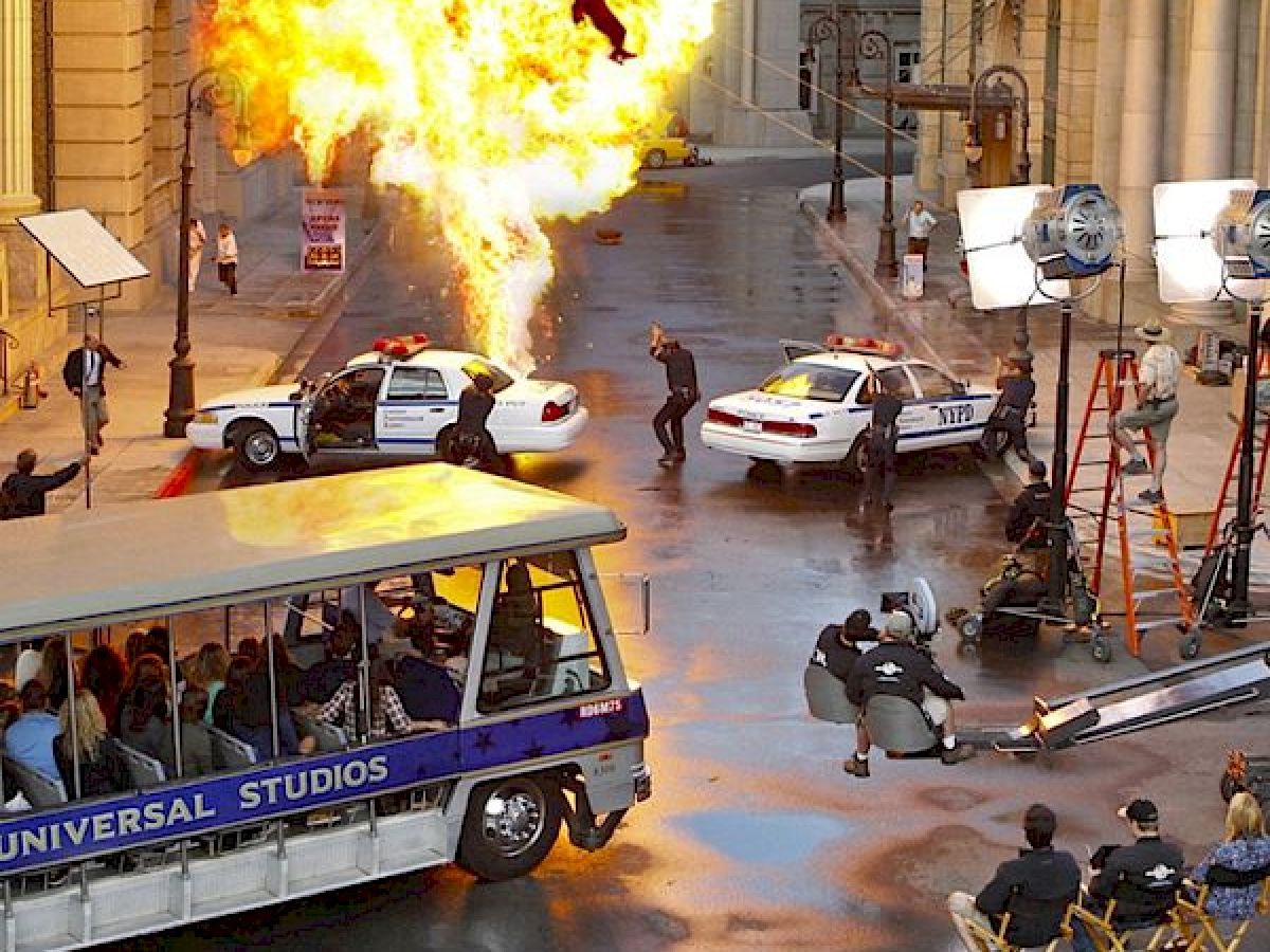 A movie scene is being filmed with an explosion, police cars, and people on set, while a Universal Studios tour tram passes by with onlookers.