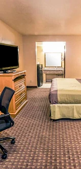 This image shows a hotel room with a bed, a flat-screen TV on a dresser, a desk chair, and an open door leading to a bathroom.