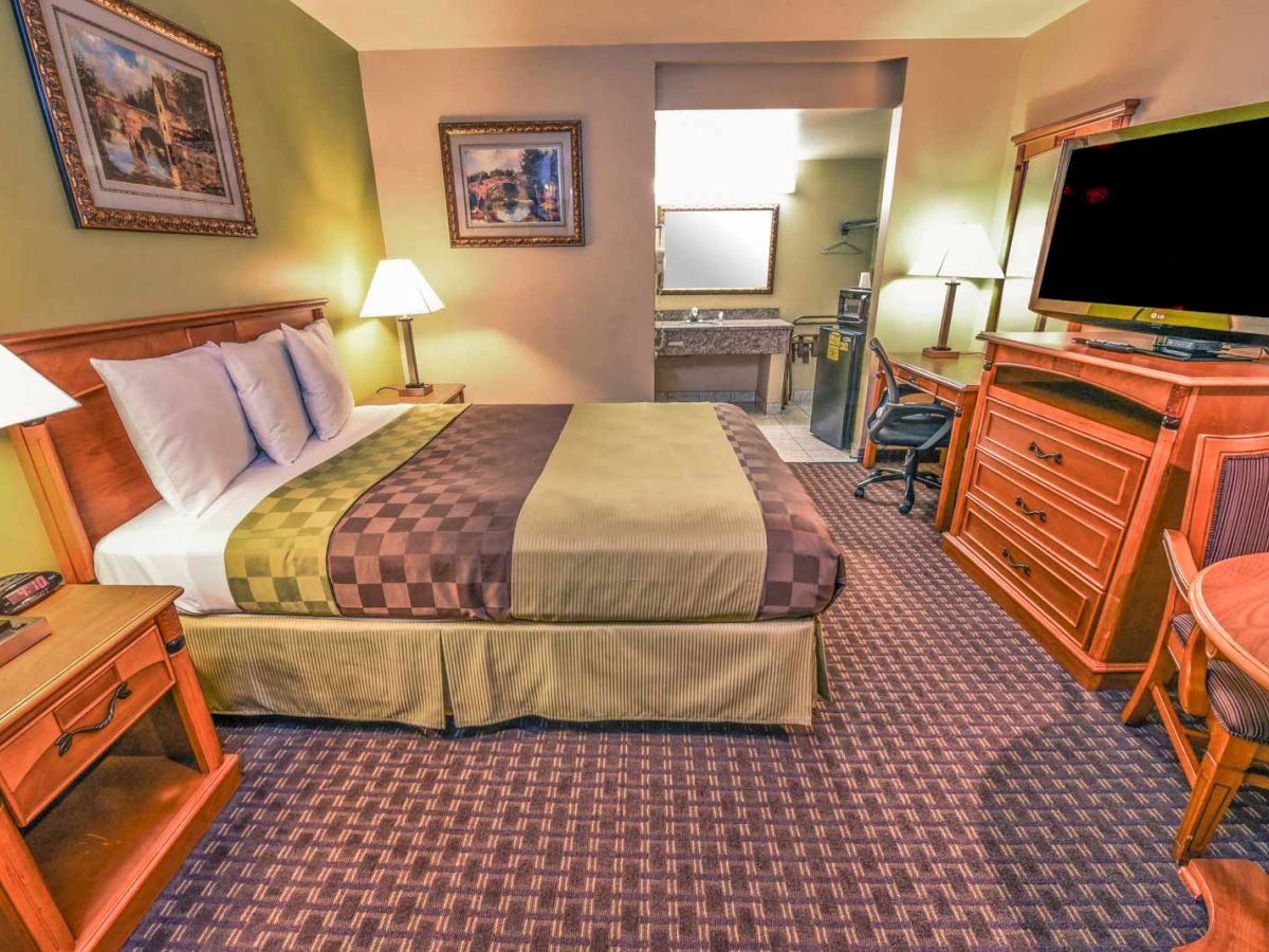 The image shows a cozy hotel room with a large bed, nightstands with lamps, a dresser with a TV, a work desk, and a sink area.