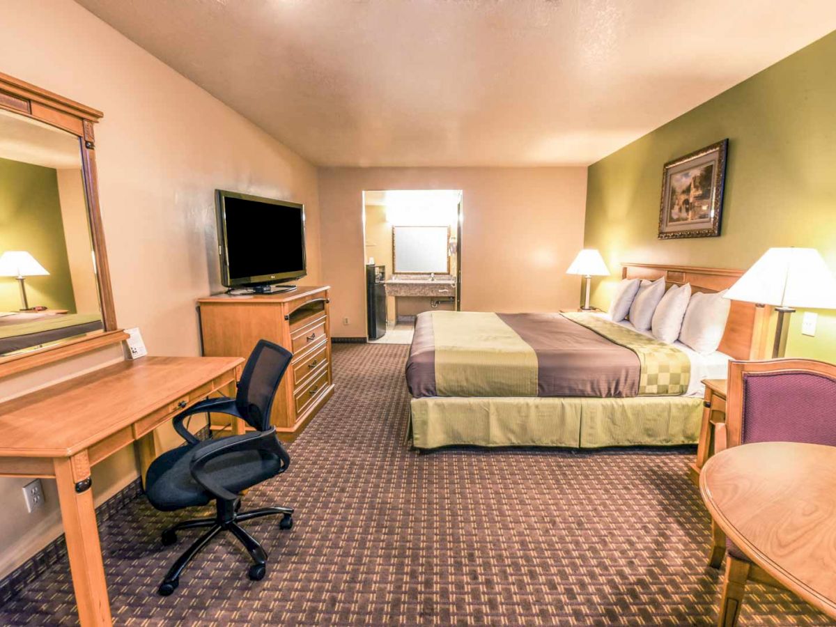 A neatly organized hotel room featuring a bed, desk, TV, chair, and lamps, with light green and beige decor and a large mirror on one wall.